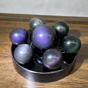 May include: Seven iridescent obsidian spheres in shades of purple, green, and black are arranged on a black, circular display stand. The spheres vary in size, with the largest in the centre. The polished surfaces reflect light, creating a shimmering effect.