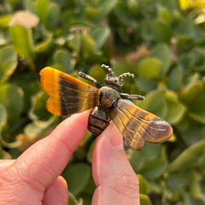May include: A detailed bee figurine with amber and brown wings, a textured body, and delicate antennae. The bee is held in a hand against a blurred green background, showcasing the intricate details of the insect sculpture.