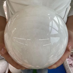 May include: A large, opaque white sphere, held in hands. The sphere has a smooth, polished surface with subtle internal veining. The light reflects off the surface, creating bright highlights. The sphere appears to be made of stone or crystal.