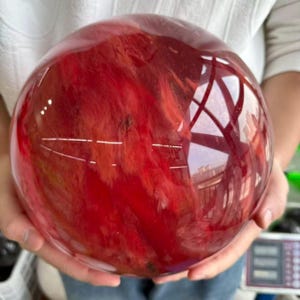 Peut inclure: Une grande sphère polie d'un rouge profond, présentant des motifs internes ressemblant à des nuages tourbillonnants. La surface réfléchit la lumière, créant un aspect brillant. La sphère est tenue dans deux mains.