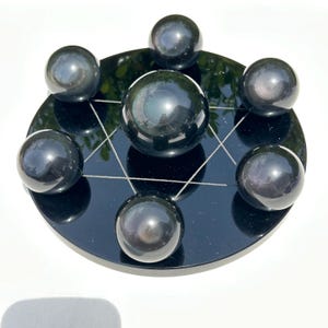 May include: A black obsidian crystal grid set. The set features seven polished obsidian spheres on a black, circular base with a white geometric design. The spheres have a reflective surface, and the base is glossy.
