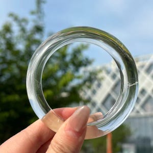 Peut inclure: Un bracelet jonc transparent et clair. Le bracelet circulaire est présenté sur un fond de feuillage vert et de ciel bleu. Le bracelet est fait d'un matériau transparent, et la lumière se reflète sur la surface.