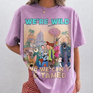 May include: A light purple short-sleeve t-shirt featuring a graphic print of various animated animal characters from Zootopia. The text WE'RE WILD AND WE CAN'T BE TAMED is printed in light blue and beige above and below the central illustration.