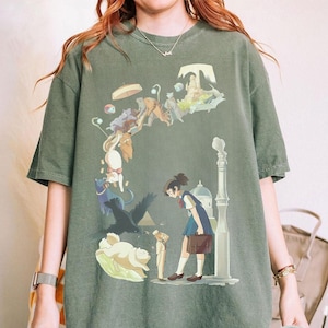 Puede incluir: Camiseta verde musgo con una ilustraci&oacute;n de estilo anime. El gr&aacute;fico representa a una chica con uniforme escolar, rodeada de gatos flotantes, un p&aacute;jaro negro y otros elementos caprichosos. Una farola y un edificio tambi&eacute;n est&aacute;n en el dise&ntilde;o.