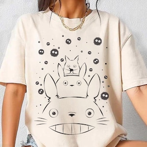 May include: Cream-colored t-shirt featuring a black and white graphic of a large, smiling cartoon character with two smaller characters on top, and several small, round, black figures scattered around the design.