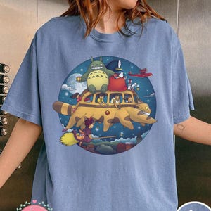 May include: A light blue t-shirt featuring a colorful graphic of a yellow bus with cartoon characters, including a large green creature, a red bag, and a small airplane. The shirt has the words "Blue Jean" printed on it.