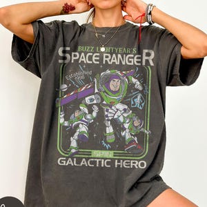 May include: Dark grey t-shirt with a graphic of Buzz Lightyear, a space ranger, and the text "Buzz Lightyear's Space Ranger" and "Galactic Hero". The design includes a green and purple spaceship and the year "Established 1998".