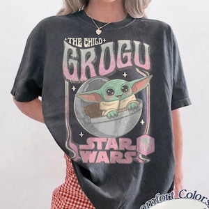 Pu&ograve; includere: Una t-shirt a maniche corte grigio antracite con una stampa grafica di Grogu di Star Wars seduto in una capsula fluttuante. Il testo recita THE CHILD, GROGU e STAR WARS in un carattere sfumato rosa e bianco, accentato da piccole stelle e icona.