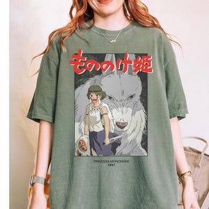 May include: An olive green t-shirt with a graphic from the animated film "Princess Mononoke." The design includes Japanese text, a girl, and a wolf. The text "PRINCESS MONONOKE 1997" is also visible.