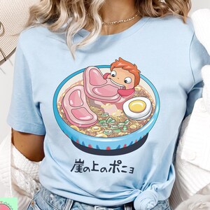 May include: Light blue t-shirt featuring a cartoon illustration of a bowl of ramen with a character holding a piece of meat. The ramen bowl is blue with a white rim and contains noodles, meat, and an egg. Japanese text is below the bowl.