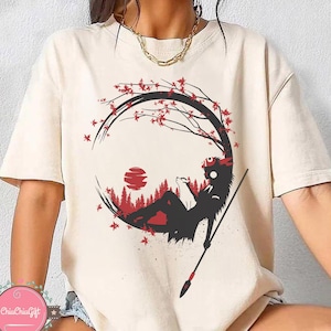 May include: Cream-colored t-shirt featuring a black and red graphic design. The design depicts a silhouette of a figure reclining within a circular frame, with a spear, a red sun, and a forest scene. The shirt has short sleeves.