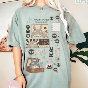 May include: A light green t-shirt featuring Studio Ghibli characters and Japanese text. The design includes illustrations of Totoro, soot sprites, and other characters from the animated film. The shirt has a relaxed fit and short sleeves.
