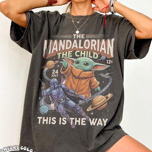 May include: A charcoal gray short-sleeve t-shirt featuring a graphic print of The Mandalorian and The Child in space. The design includes planets, stars, and the text THE MANDALORIAN, THE CHILD, 24 JULY, 12c, and THIS IS THE WAY in a vintage style.