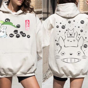 May include: Cream-colored hoodie featuring illustrations from a Japanese animated film. The front displays a white rabbit-like creature with black soot sprites and Japanese text. The back shows a large character with smaller characters.