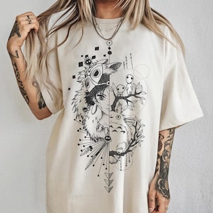 Puede incluir: Camiseta color crema con un dise&ntilde;o gr&aacute;fico en blanco y negro. El dise&ntilde;o est&aacute; dividido verticalmente, mostrando un personaje con una m&aacute;scara en un lado y una escena de bosque con figuras en el otro. Formas geom&eacute;tricas y flechas completan el dise&ntilde;o.