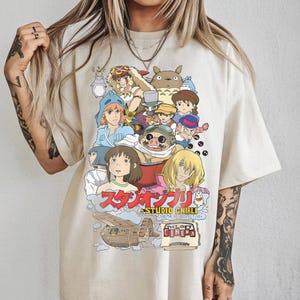 May include: Cream-colored t-shirt featuring a colorful graphic of Studio Ghibli characters. The design includes Japanese text and the words "THE LOFT CINEMA RETROSPECTIVE". The shirt has a relaxed fit and short sleeves.