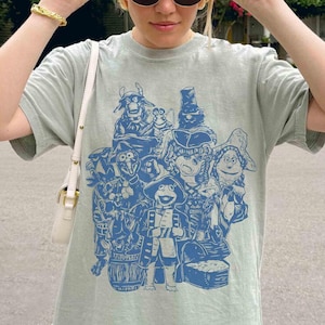May include: A light green t-shirt featuring a blue illustration of various cartoon characters. The design includes a variety of whimsical figures, some wearing hats and costumes. The shirt has short sleeves and a crew neck.