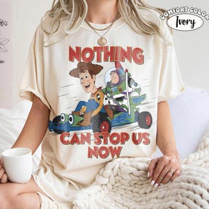 May include: An ivory t-shirt featuring a graphic of Woody and Buzz Lightyear riding a blue race car. The text above reads NOTHING and below reads CAN STOP US NOW. A circular badge in the top right corner says COMFORT COLOR IVORY.