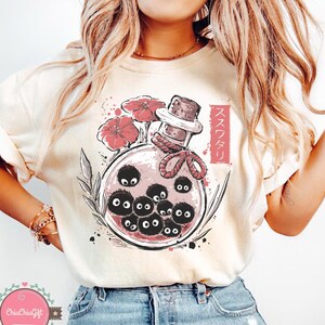 Puede incluir: Camiseta color crema con una ilustraci&oacute;n caprichosa de una botella de poci&oacute;n. Dentro de la botella hay varias criaturas negras y redondas con ojos grandes. Flores rojas y texto japon&eacute;s acent&uacute;an el dise&ntilde;o. La botella tiene un tap&oacute;n de corcho y est&aacute; atada con una cuerda roja.