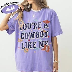 K&ouml;nnte beinhalten: Ein violettes Kurzarm-T-Shirt mit dem Text "YOU'RE A COWBOY LIKE ME" in dunkelblauen Buchstaben. Die Grafik zeigt einen braunen Cowboyhut, eine Rakete, einen Sheriffstern, einen Ball und braune Cowboystiefel, akzentuiert durch kleine Sterne und Wolken.