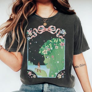 May include: Dark gray t-shirt featuring a graphic of a green-skinned woman with a floral crown and a pink ribbon. The design includes a landscape with a figure and a dog. The shirt has short sleeves.