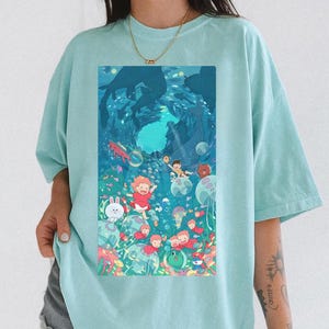 K&ouml;nnte beinhalten: Hellblaues T-Shirt mit einer farbenfrohen Illustration einer Unterwasserszene mit Zeichentrickfiguren, Fischen und Blasen. Das grafische Design ist mittig platziert und zeigt eine lebendige, skurrile Unterwasserwelt.