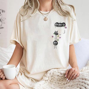 May include: Cream-colored t-shirt featuring a pocket design with cartoon soot sprites. The pocket contains three soot sprites, while two more are below, with colorful star accents. The shirt has short sleeves and a relaxed fit.