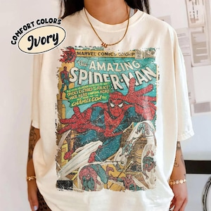 K&ouml;nnte beinhalten: Ein elfenbeinfarbenes T-Shirt mit einer Vintage-Marvel-Comics-Grafik von The Amazing Spider-Man. Der Druck zeigt Spider-Man in seinem rot-blauen Anzug, mit Comic-Text, einschlie&szlig;lich "The Amazing Spider-Man" und "Chameleon" in einem abgenutzten Stil.