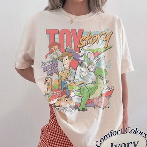 May include: An ivory short-sleeved t-shirt featuring a large graphic print of Toy Story characters including Woody, Buzz Lightyear, Jessie, Rex, Slinky Dog, Bullseye, and Hamm. The text reads Toy Story, Jessie SINCE 1995, Woody, Buzz, Slinky Dog, and Bullseye.