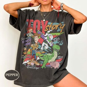 May include: A charcoal gray short-sleeve t-shirt featuring a graphic print of Toy Story characters including Woody, Buzz Lightyear, Jessie, Rex, Slinky Dog, and Bullseye. The text reads Toy Story, Jessie Since 1995, Woody, Buzz, Rex, and Bullseye.