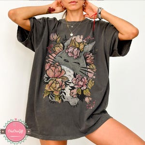May include: Dark grey t-shirt featuring a floral cat graphic. The design includes a grey cat face surrounded by pink and yellow flowers. The shirt has short sleeves and a relaxed fit, suitable for casual wear.