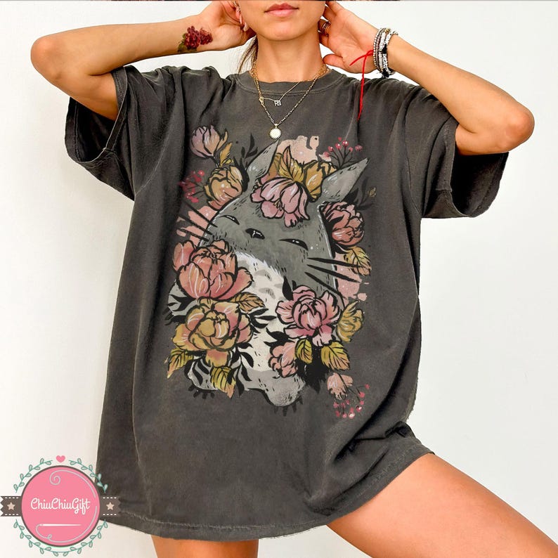 May include: Dark grey t-shirt featuring a floral cat graphic. The design includes a grey cat face surrounded by pink and yellow flowers. The shirt has short sleeves and a relaxed fit, suitable for casual wear.