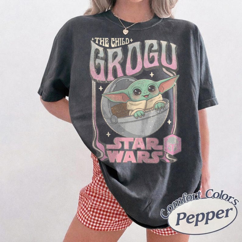 Pu&ograve; includere: Una t-shirt a maniche corte grigio antracite con una stampa grafica di Grogu di Star Wars seduto in una capsula fluttuante. Il testo recita THE CHILD, GROGU e STAR WARS in un carattere sfumato rosa e bianco, accentato da piccole stelle e icona.