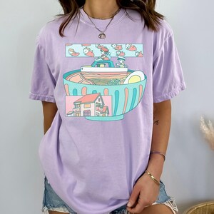 Puede incluir: Camiseta lila con una ilustraci&oacute;n caprichosa de un cuenco de ramen. El cuenco est&aacute; lleno de fideos, un huevo y otros ingredientes. Encima del cuenco, un dise&ntilde;o rectangular muestra personajes de dibujos animados y peces. Debajo hay una casa.