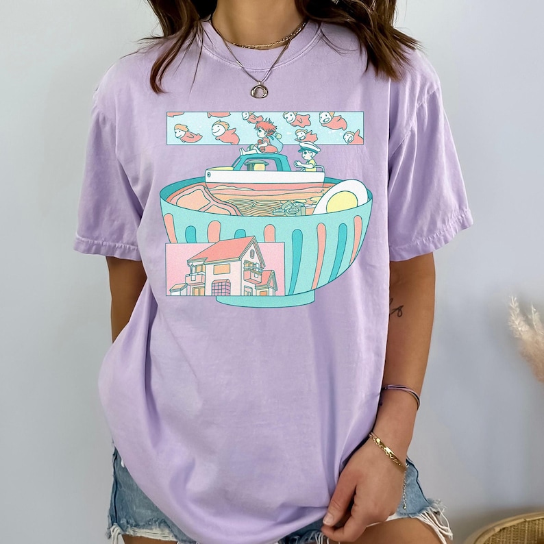 Puede incluir: Camiseta lila con una ilustraci&oacute;n caprichosa de un cuenco de ramen. El cuenco est&aacute; lleno de fideos, un huevo y otros ingredientes. Encima del cuenco, un dise&ntilde;o rectangular muestra personajes de dibujos animados y peces. Debajo hay una casa.
