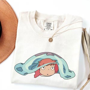 May include: Cream-colored t-shirt featuring a cartoon character with red hair peeking out from under a blue blanket. A gold necklace with a pendant rests on the shirt. The shirt has a Comfort Colors tag.