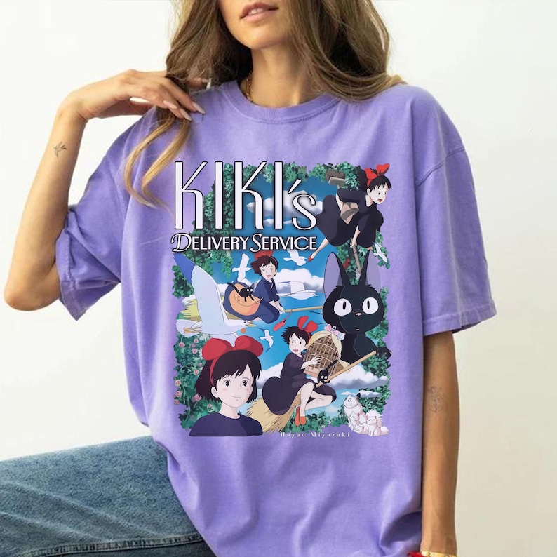 May include: A lilac-colored t-shirt featuring a graphic from "Kiki's Delivery Service." The design includes the text "Kiki's Delivery Service" and illustrations of Kiki, Jiji the cat, and other characters from the animated film.
