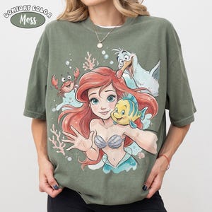 Puede incluir: Una camiseta de manga corta color verde musgo con un estampado gr&aacute;fico de una sirena pelirroja, un pez amarillo, un peque&ntilde;o cangrejo rojo y una gaviota blanca. El dise&ntilde;o incluye detalles de coral y burbujas. Etiqueta Comfort Color Moss.