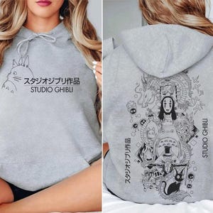 May include: A light gray hoodie featuring a black graphic of a cartoon character and Japanese text. The front of the hoodie has the text "STUDIO GHIBLI". The back of the hoodie has a larger graphic with multiple characters and the same text.