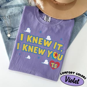 May include: A violet short-sleeved t-shirt featuring the yellow text I KNEW IT, I KNEW YOU with a colourful ball graphic and a red heart containing the letters TS. The design includes small white cloud and blue star icons printed on the fabric.