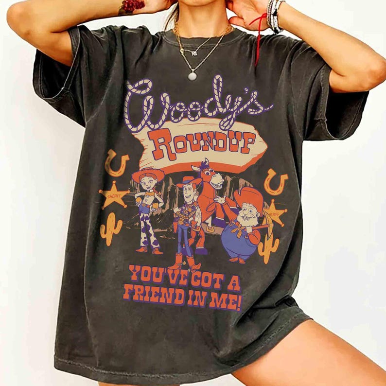 May include: Dark gray t-shirt featuring a colorful graphic of Woody's Roundup characters. The design includes the text "Woody's Roundup" and "You've Got a Friend in Me!" with cowboy-themed illustrations.