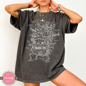 May include: Dark grey t-shirt featuring a detailed white blueprint design of a fantastical moving structure. The design includes labeled components like "wind gauge" and "stabilizer legs." The shirt has a relaxed fit.