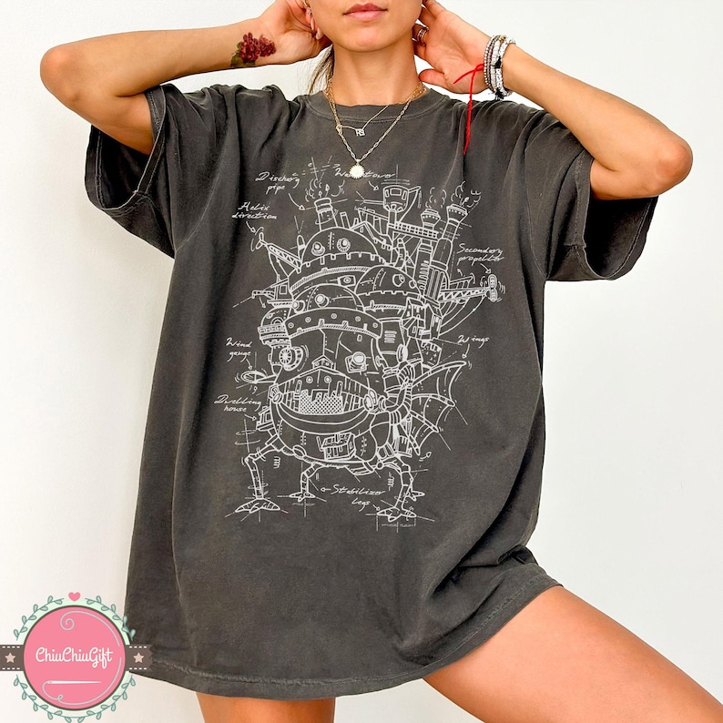 May include: Dark grey t-shirt featuring a detailed white blueprint design of a fantastical moving structure. The design includes labeled components like "wind gauge" and "stabilizer legs." The shirt has a relaxed fit.