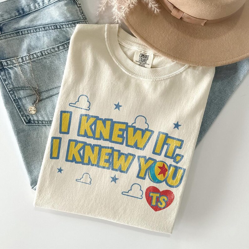 May include: A cream-coloured short-sleeve t-shirt featuring the blue and yellow text I KNEW IT, I KNEW YOU with a red heart containing the letters TS, small blue stars, and cloud outlines. The shirt has a crew neckline and a Comfort Colors brand label.