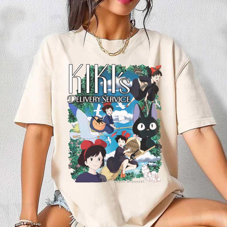 May include: Beige t-shirt featuring a colorful graphic from Kiki's Delivery Service. The design includes the text "Kiki's Delivery Service" and illustrations of characters and scenes from the animated film. The shirt has short sleeves and a crew neck.