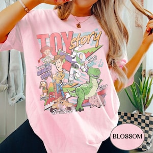 May include: A light pink t-shirt featuring a graphic print of Toy Story characters including Woody, Buzz Lightyear, Jessie, Rex, Slinky Dog, Bullseye, and Hamm. The text reads Toy Story, Jessie, Buzz, Woody, Slinky Dog, Bullseye, and Rex.