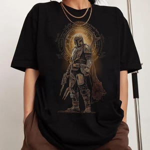 Pu&ograve; includere: Una t-shirt nera a maniche corte con una stampa grafica di un personaggio corazzato con elmo e mantello, che impugna un'arma. Il design include un emblema circolare dorato con intricati motivi testuali attorno alla figura centrale.