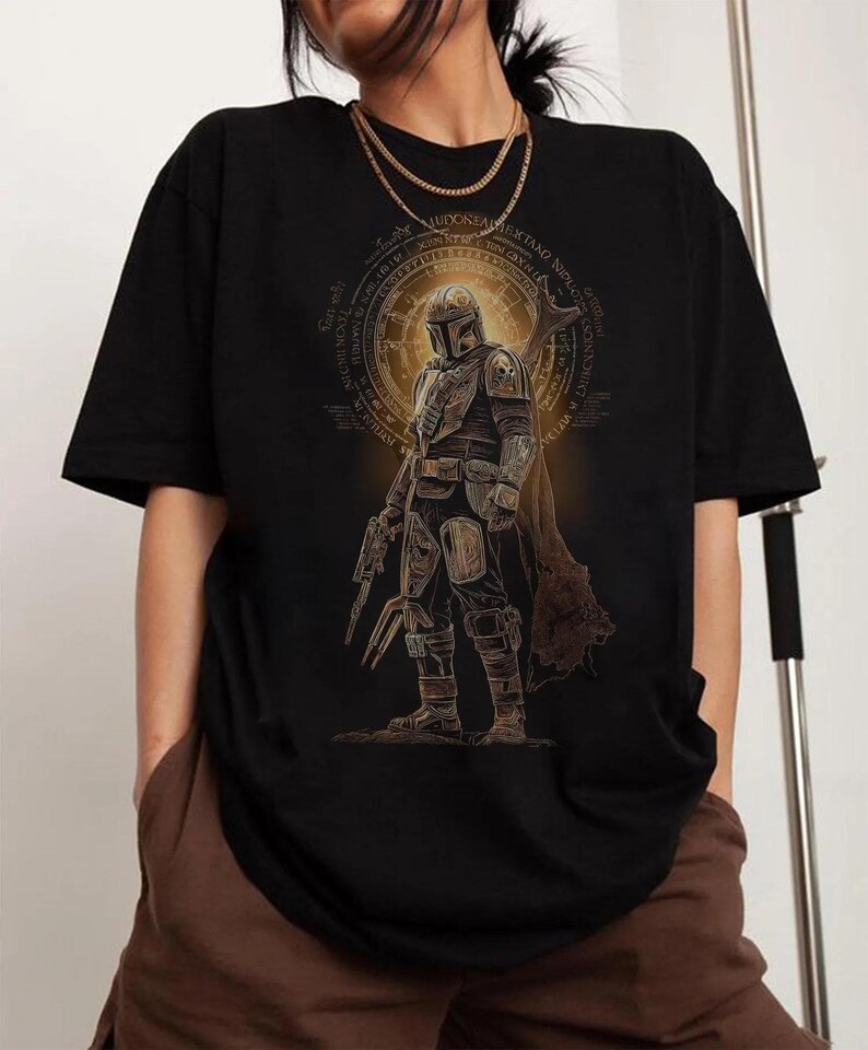 Pu&ograve; includere: Una t-shirt nera a maniche corte con una stampa grafica di un personaggio corazzato con elmo e mantello, che impugna un'arma. Il design include un emblema circolare dorato con intricati motivi testuali attorno alla figura centrale.