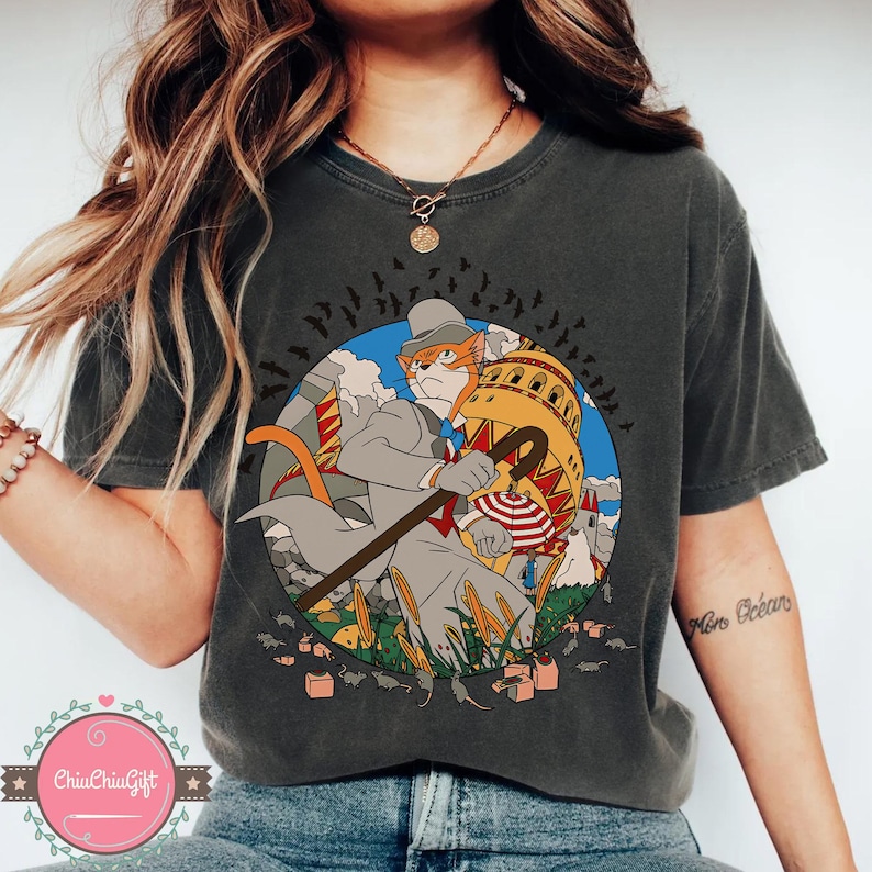 May include: Dark gray t-shirt featuring a cartoon cat in a suit, hat, and cane, set within a circular design. The cat is holding a cane and standing in front of a building. The design includes bats, grass, and small buildings.