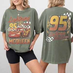 May include: An olive green short-sleeve t-shirt featuring a graphic of the red race car Lightning McQueen on the front with the text Lightning 95 MCQUEEN. The back displays the number 95 in large yellow font with flames and a checkered flag pattern.